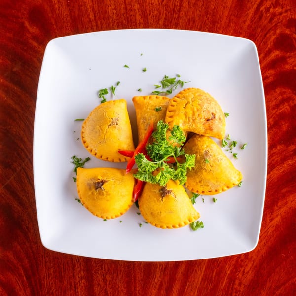 A white square plate holds six golden Jamaican patties garnished with parsley and red peppers.