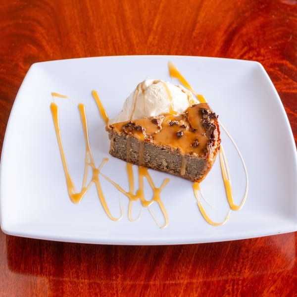 A slice of Jamaican sweet potato pudding with ice cream and caramel drizzle on a white plate.