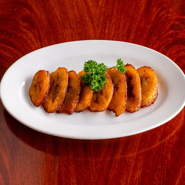 A white oval plate holds a row of golden-brown fried plantain slices garnished with fresh parsley.